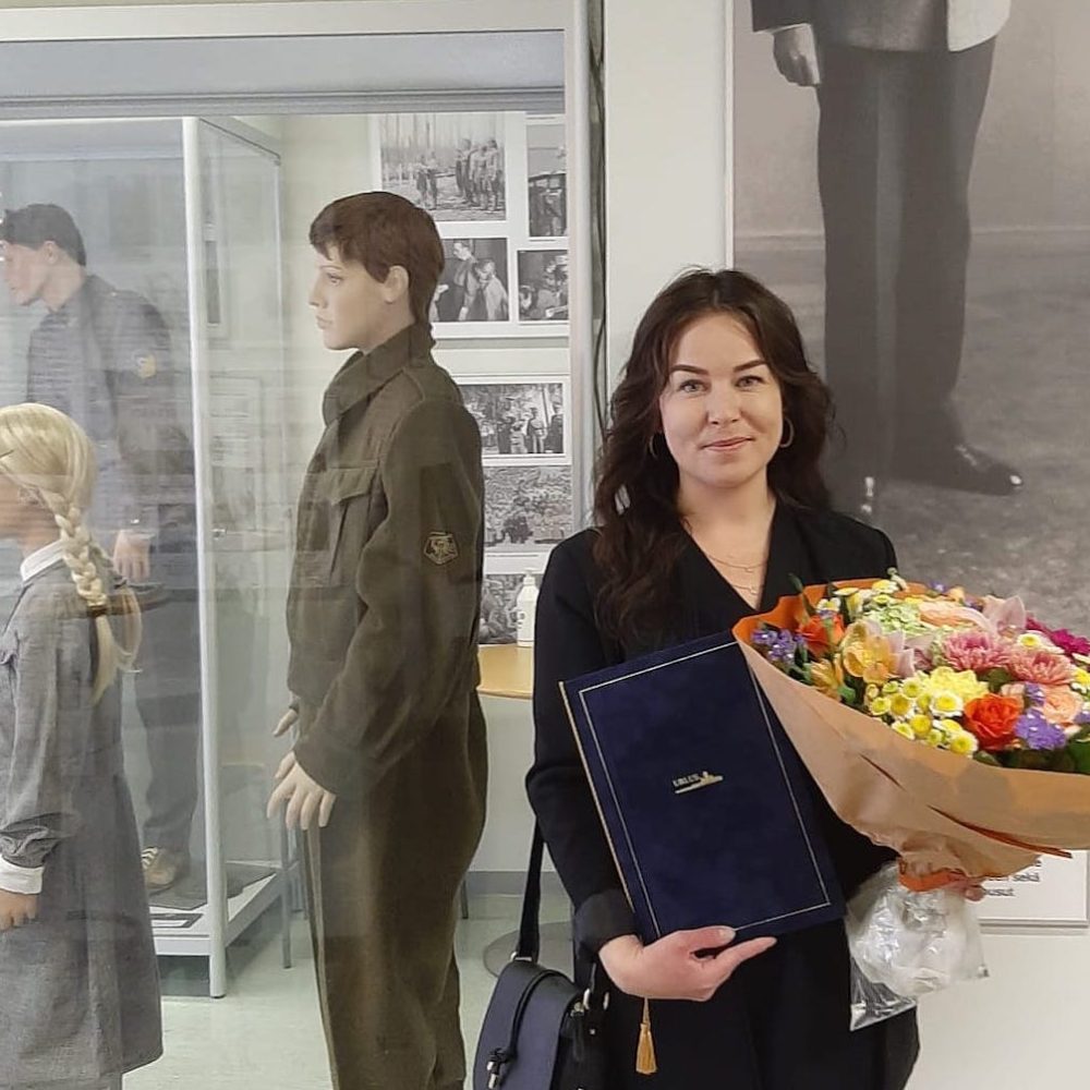 Nainen seisoo Urlus-Säätiön tiloissa suuri kukkakimppu ja kunniakirja kädessään.
