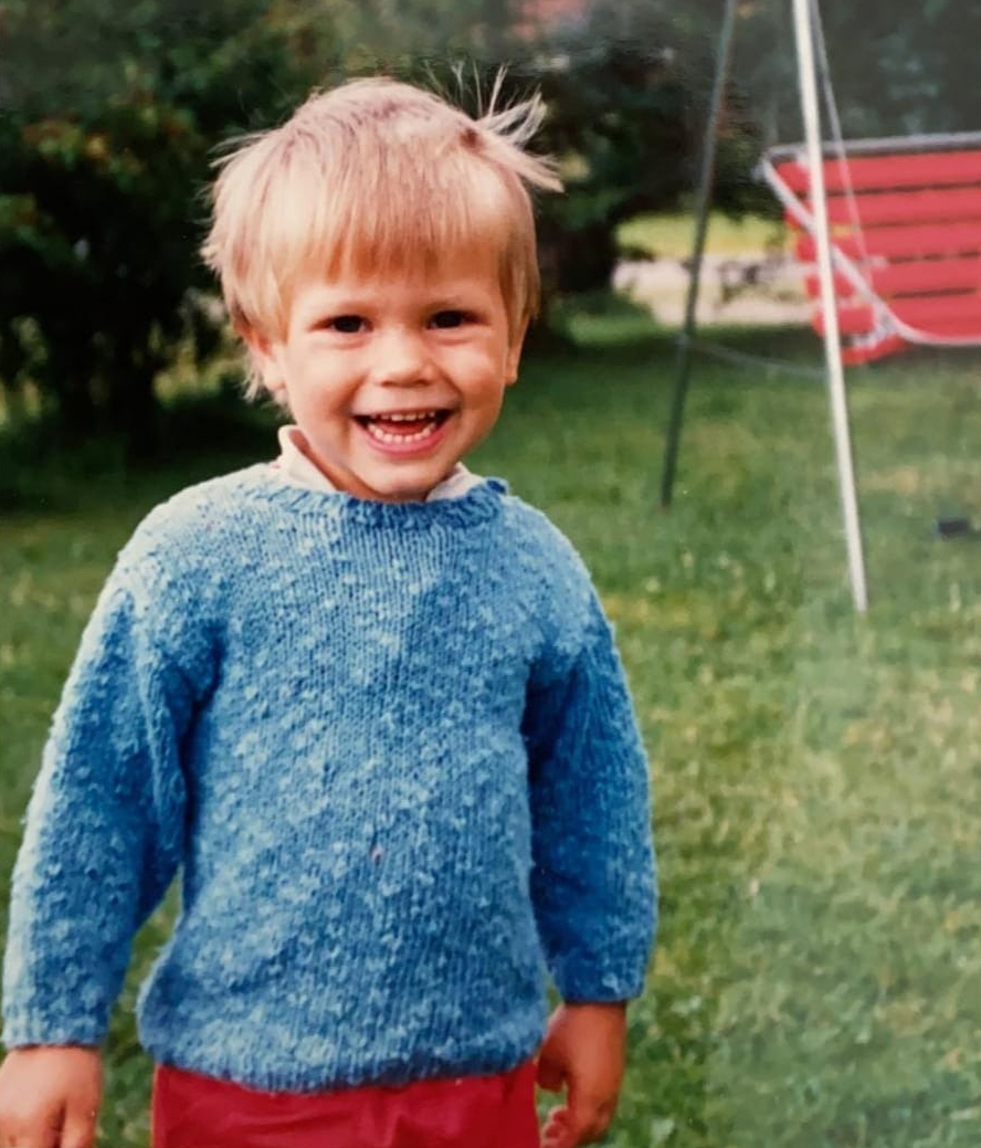 Tomas Luoto lapsena. Hän hymyilee kuvassa sininen villapaita ja punaiset housut jalassa. Taustalla näkyy nurmikko ja punainen pihakeinu.