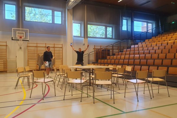 Koulun liikuntasali, jossa tyhjiä tuoleja ja pöytiä, taustalla kaksi ihmistä.