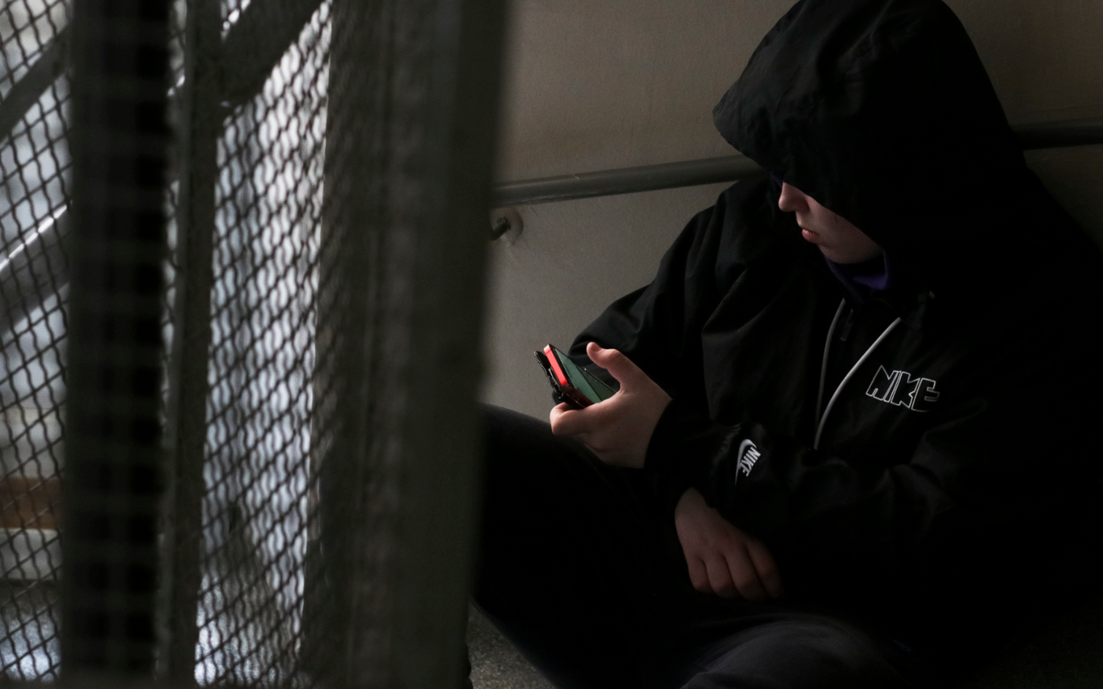 Nuori istuu rappukäytävässä selaamassa kännykkään huppu päässä. Kasvoja ei näy.