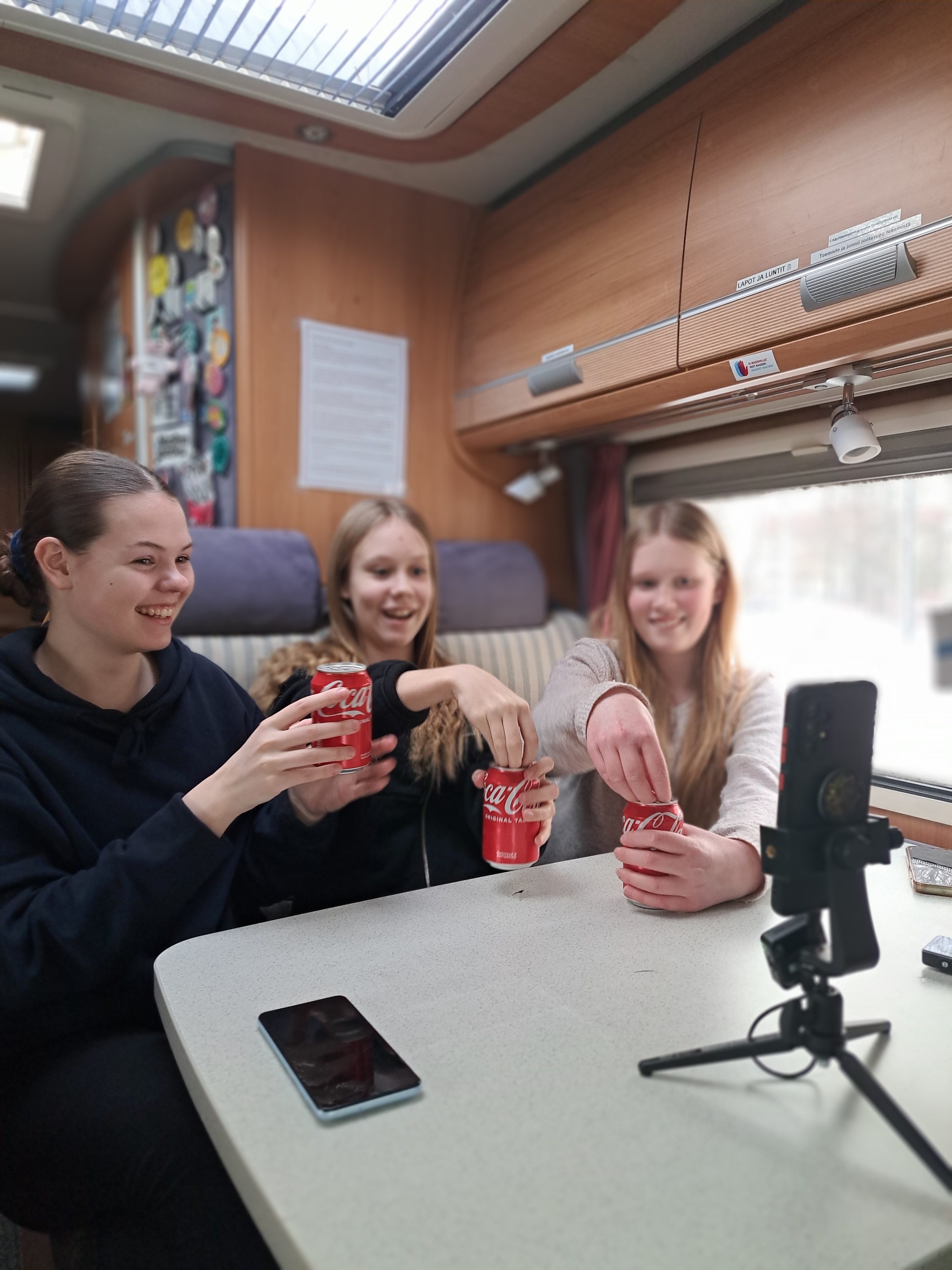 Kolme nuorta istuu Wautossa ja kuvaa kännykällä videota, jossa juovat coca-kolaa.