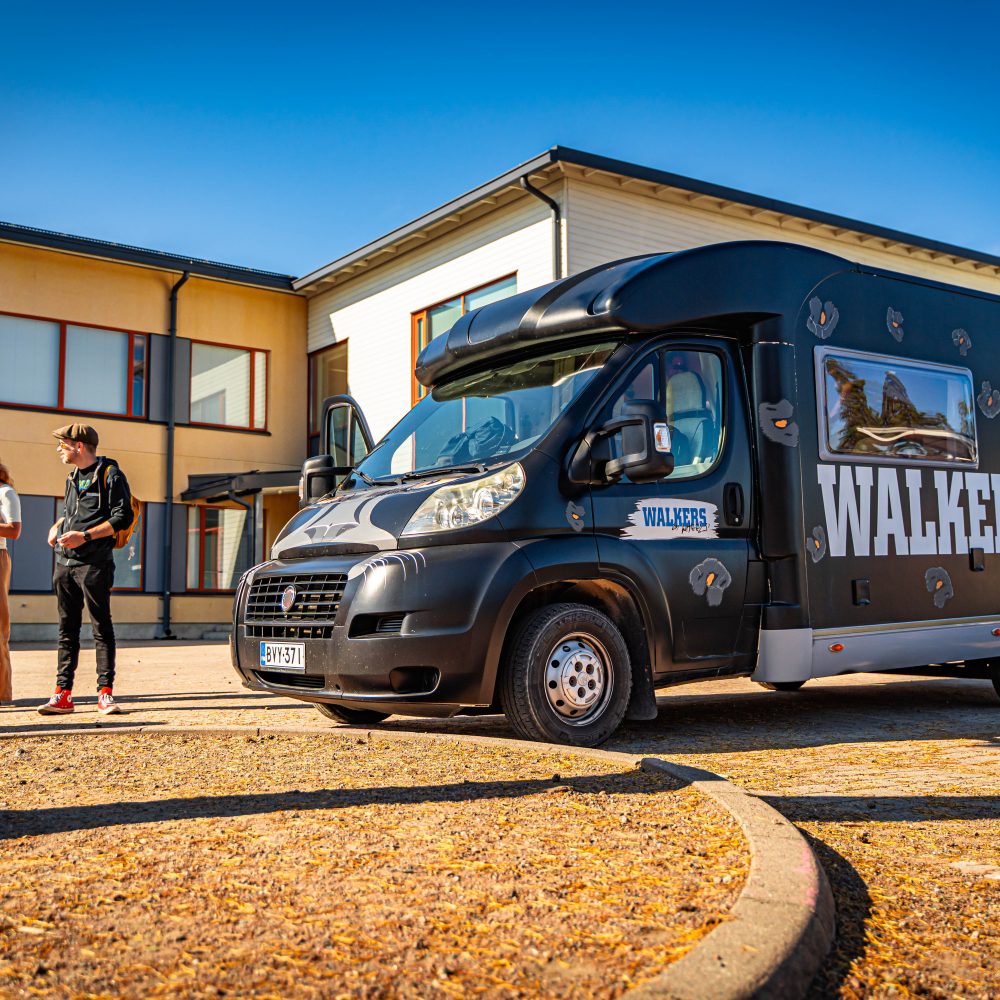 Walkers-auto aurinkoisella säällä, taustalla rakennus ja auton vieressä muutama ihminen.