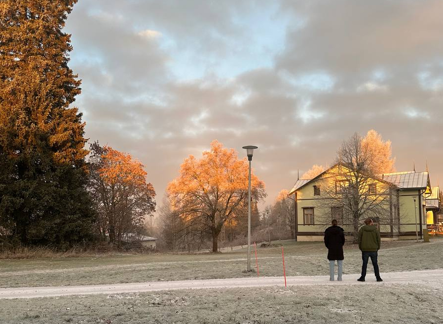 Kaksi henkilöä seisoo ulkona selkä kameraan päin, maa on huurteessa ja aurinko värjää kauempana olevien puiden latvoja.
